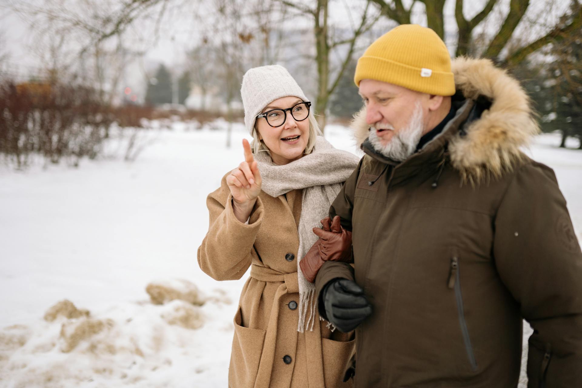 Senior couple walking in snowy park, dressed warmly in winter coats and hats, sharing a joyful moment.