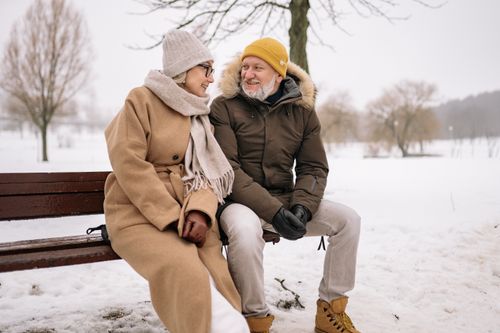 Free Happy elderly couple sitting on a snowy bench, wrapped in winter coats and sharing a joyful moment. Stock Photo