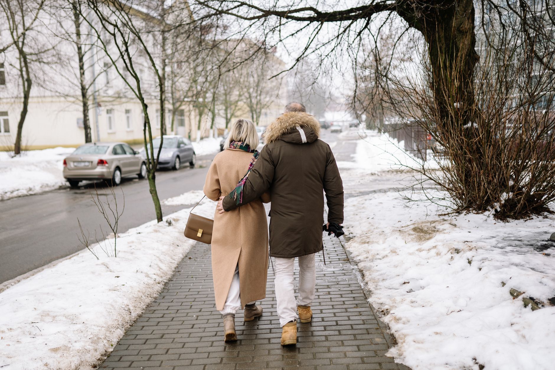 Elderly couple holding hands, walking on a snowy street in winter attire.