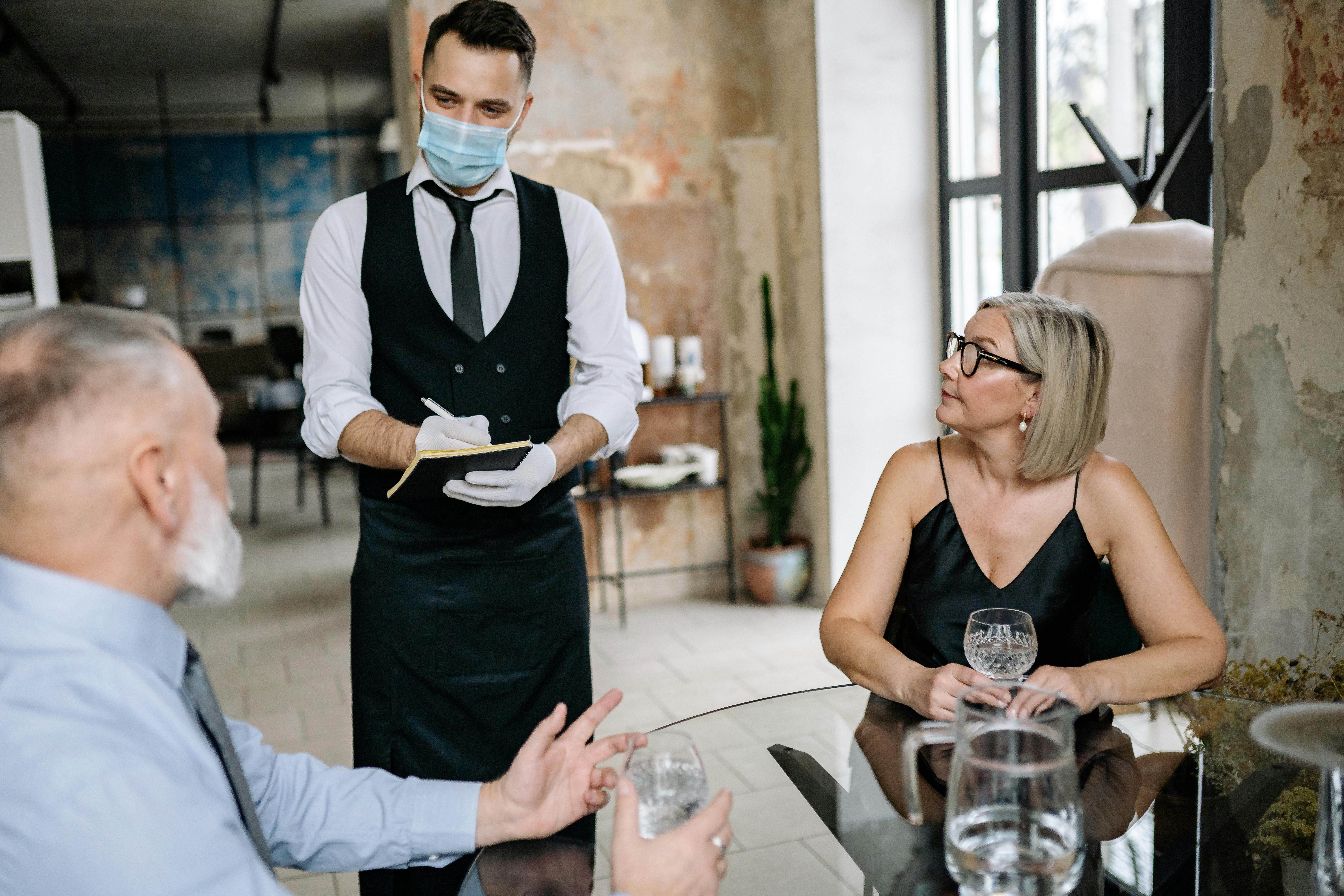 Masked waiter serves customers in a contemporary restaurant, ensuring health safety.