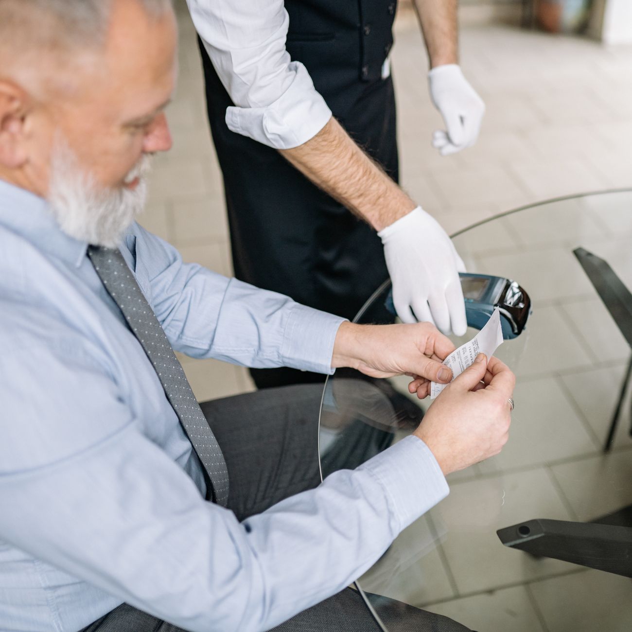 Senior man making a payment via credit card with a waiter in gloves assisting.