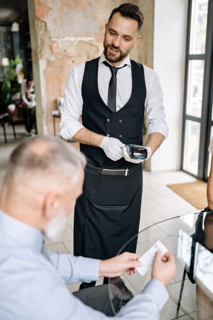 A waiter processes payment using a terminal while a customer reviews the bill, showcasing excellent service. A waiter processes payment using a terminal while a customer reviews the bill, showcasing excellent service.