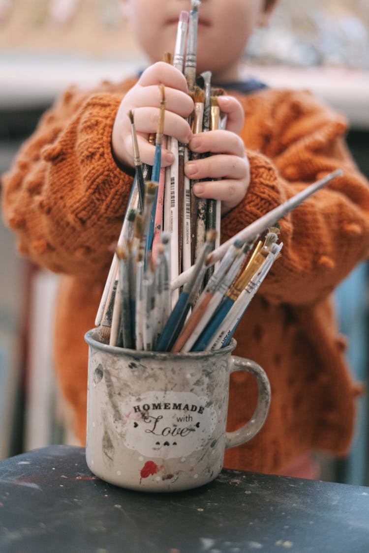 A Child Holding Paintbrushes In A Mug