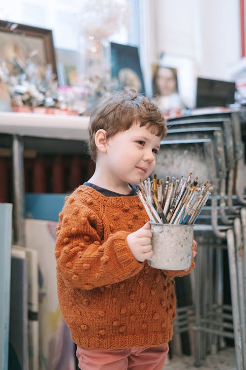 A Child in a Knitted Sweater Holding a Cup of Paintbrushes