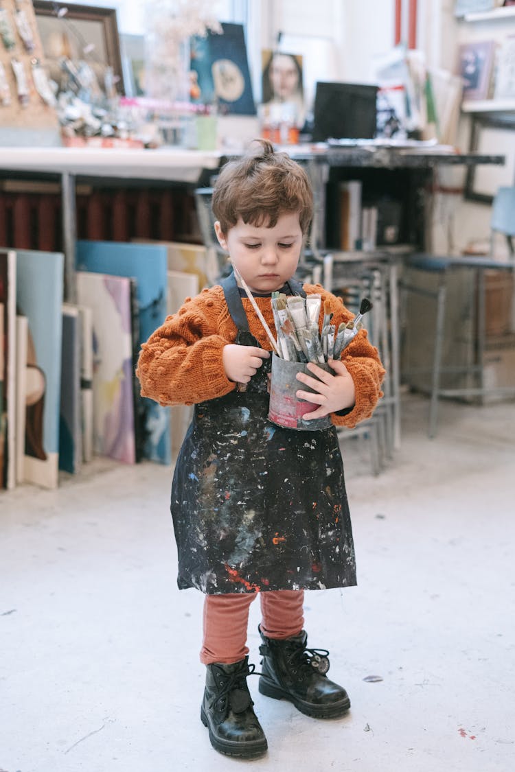 A Cute Kid Wearing Apron While Holding A Holder With Paintbrushes