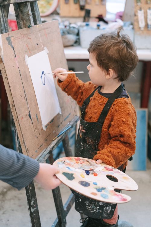 Free A young child paints with a brush on an easel, surrounded by art supplies in a classroom setting. Stock Photo