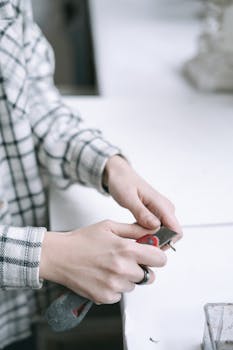 Close-up of hands sharpening a pencil using a cutter, wearing a plaid shirt indoors.