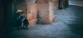 一位戴着传统帽子的老妇人坐在砖墙旁，描绘出街道的孤独场景