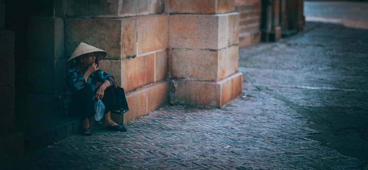 一位戴着传统帽子的老妇人坐在砖墙旁，描绘出街道的孤独场景