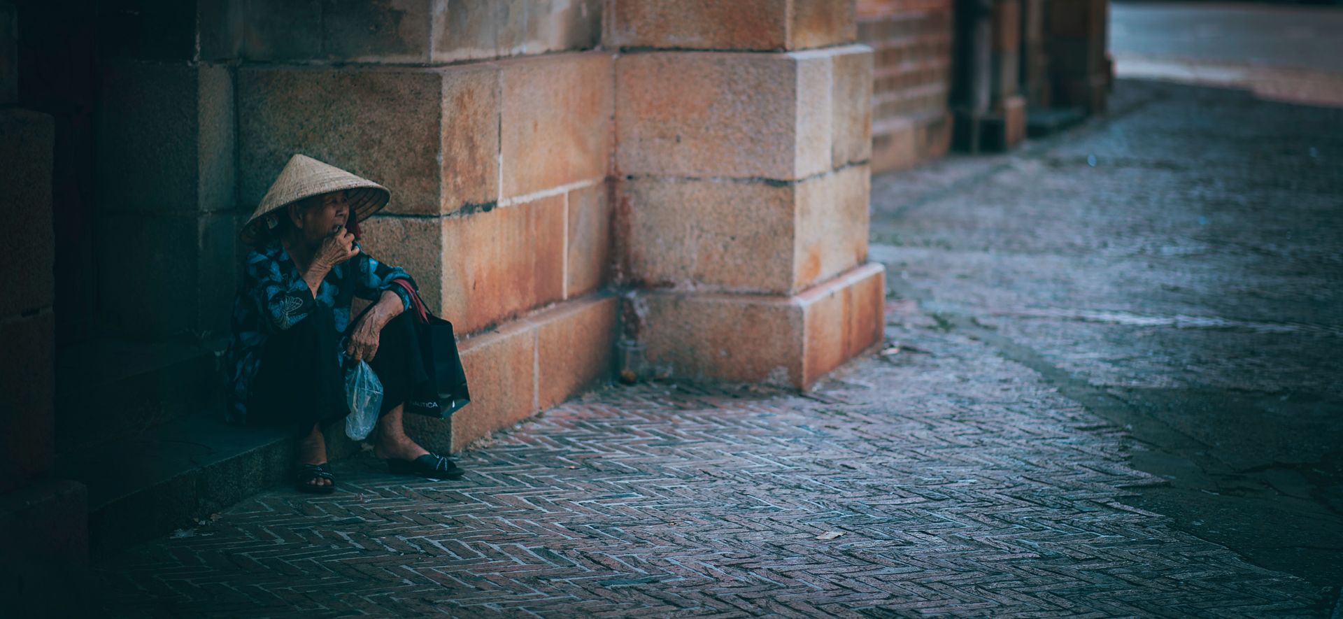 Elderly woman in traditional hat sitting by a brick wall, a street scene depicting solitude.
