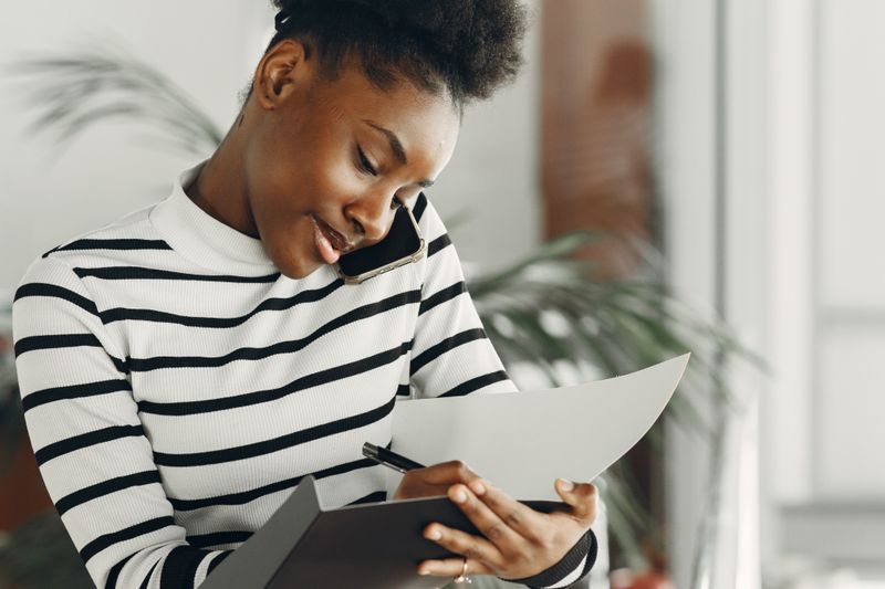 A Black woman in a black and white striped turtleneck holds a phone to her ear with her shoulder while writing notes on a page in an open folder, standing in a bright room with a houseplant behind her.