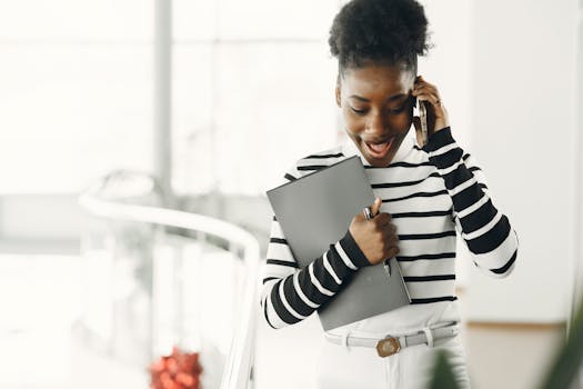 A smiling young woman with curly hair engages in a phone call while holding a notebook indoors.
