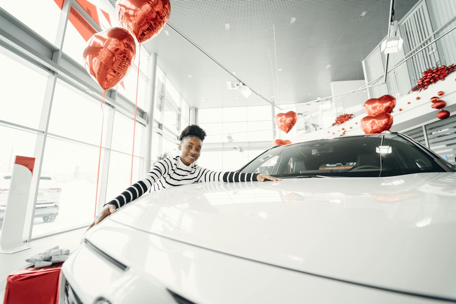 Joyful woman celebrating new car purchase at dealership with heart balloons.