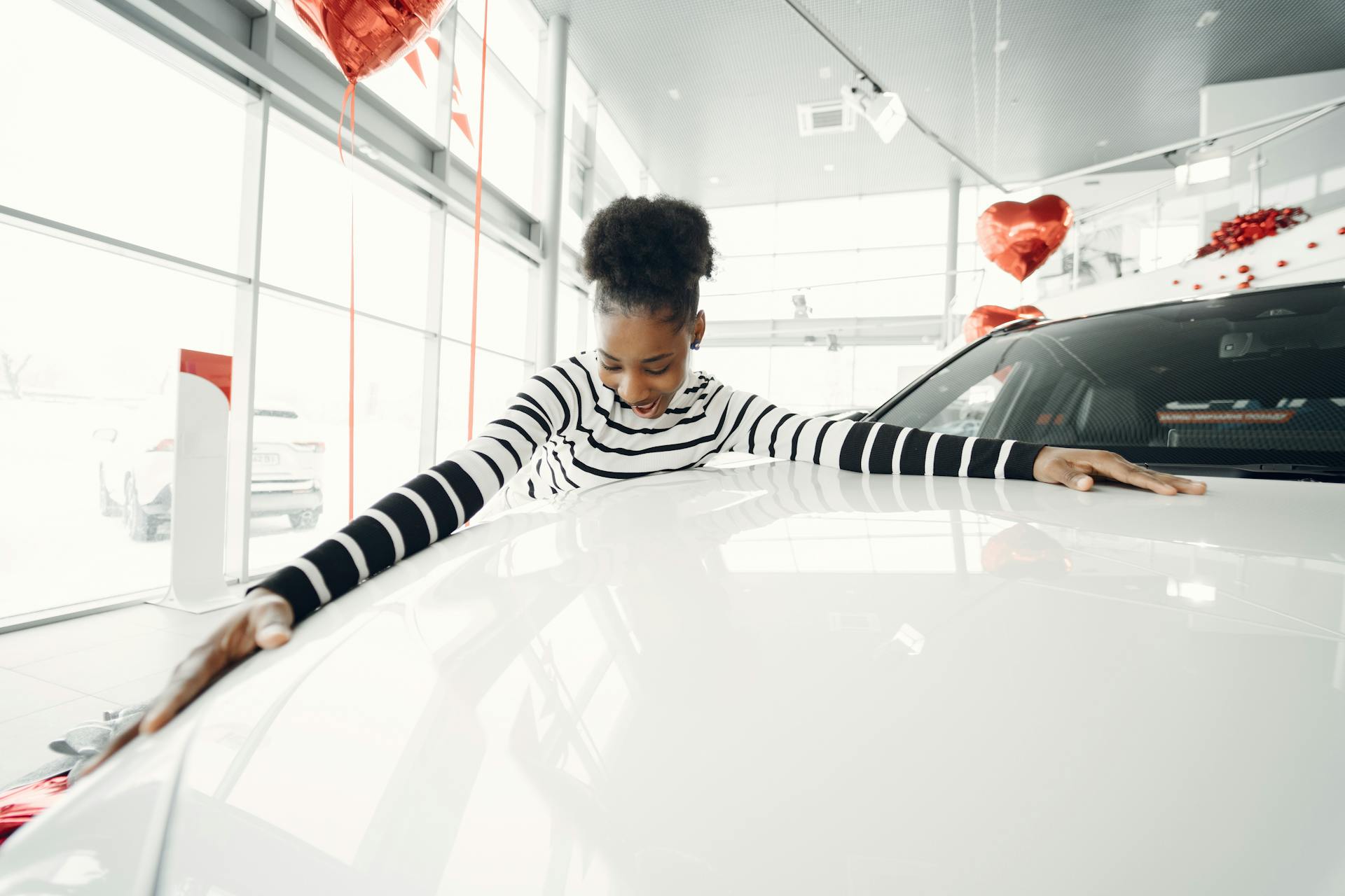 African American woman joyfully embracing a car in a dealership with heart-shaped balloons.