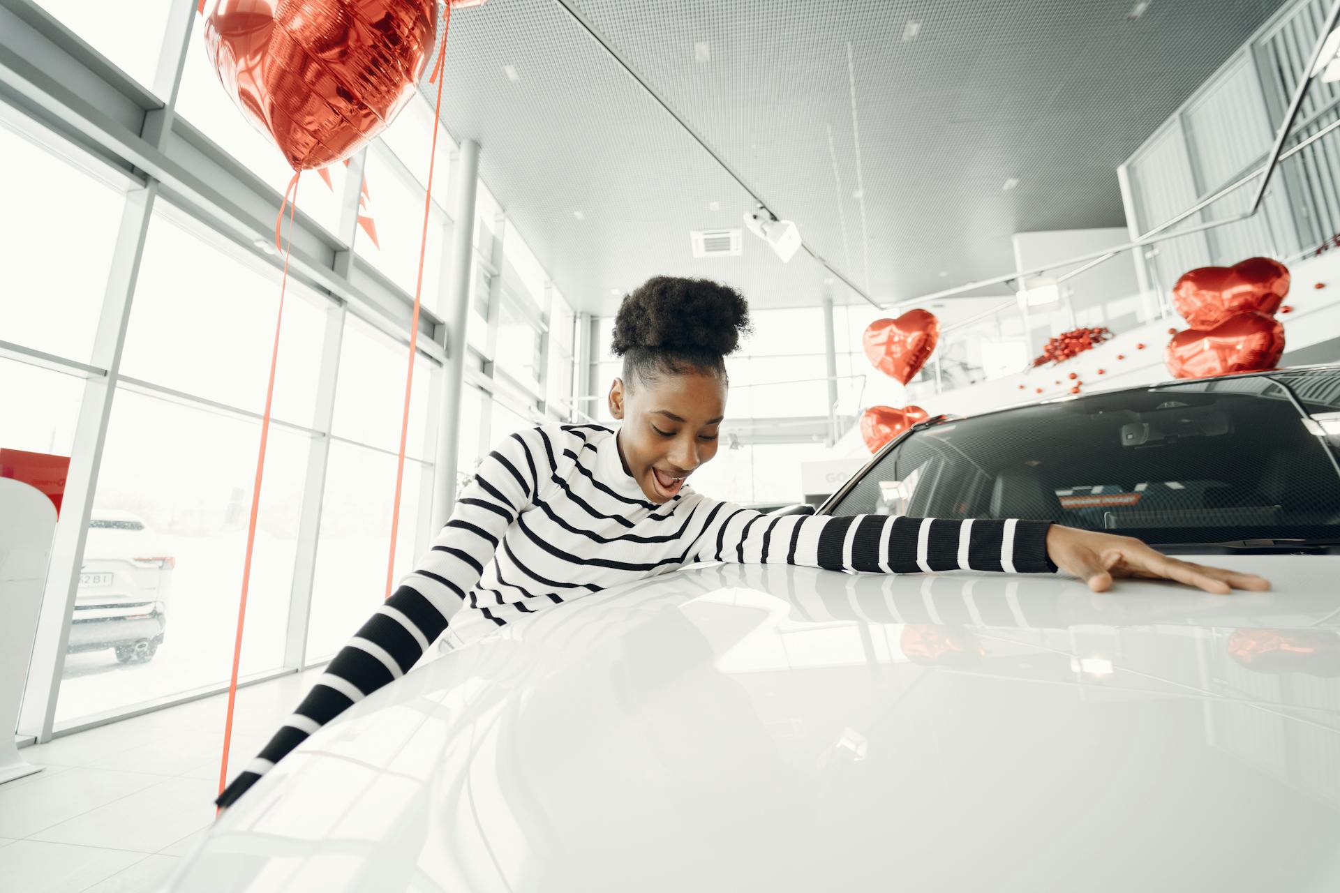 Excited woman embraces her new car at a dealership, surrounded by red balloons.