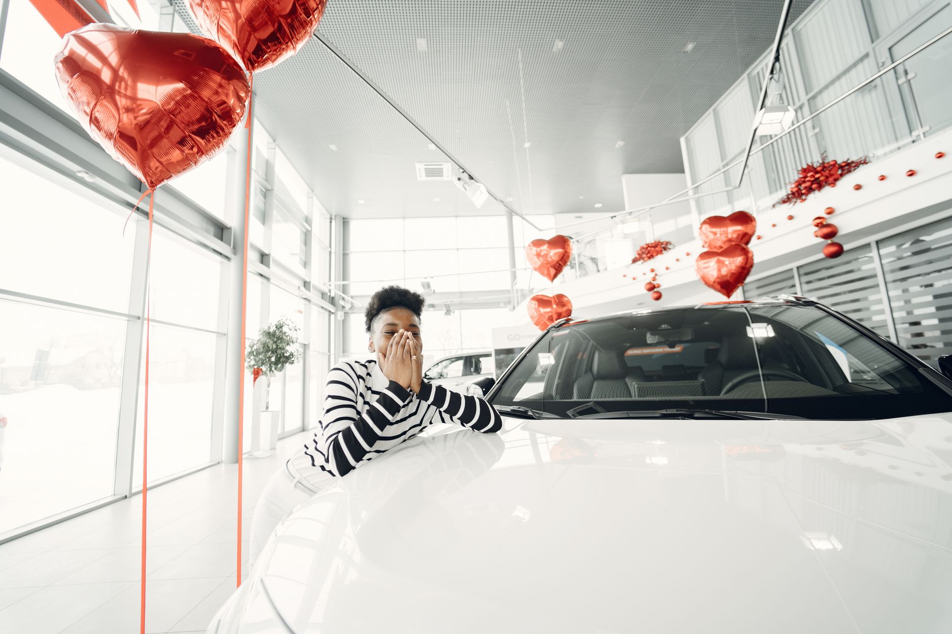 A young woman expresses excitement at a car dealership, surrounded by heart-shaped red balloons.
