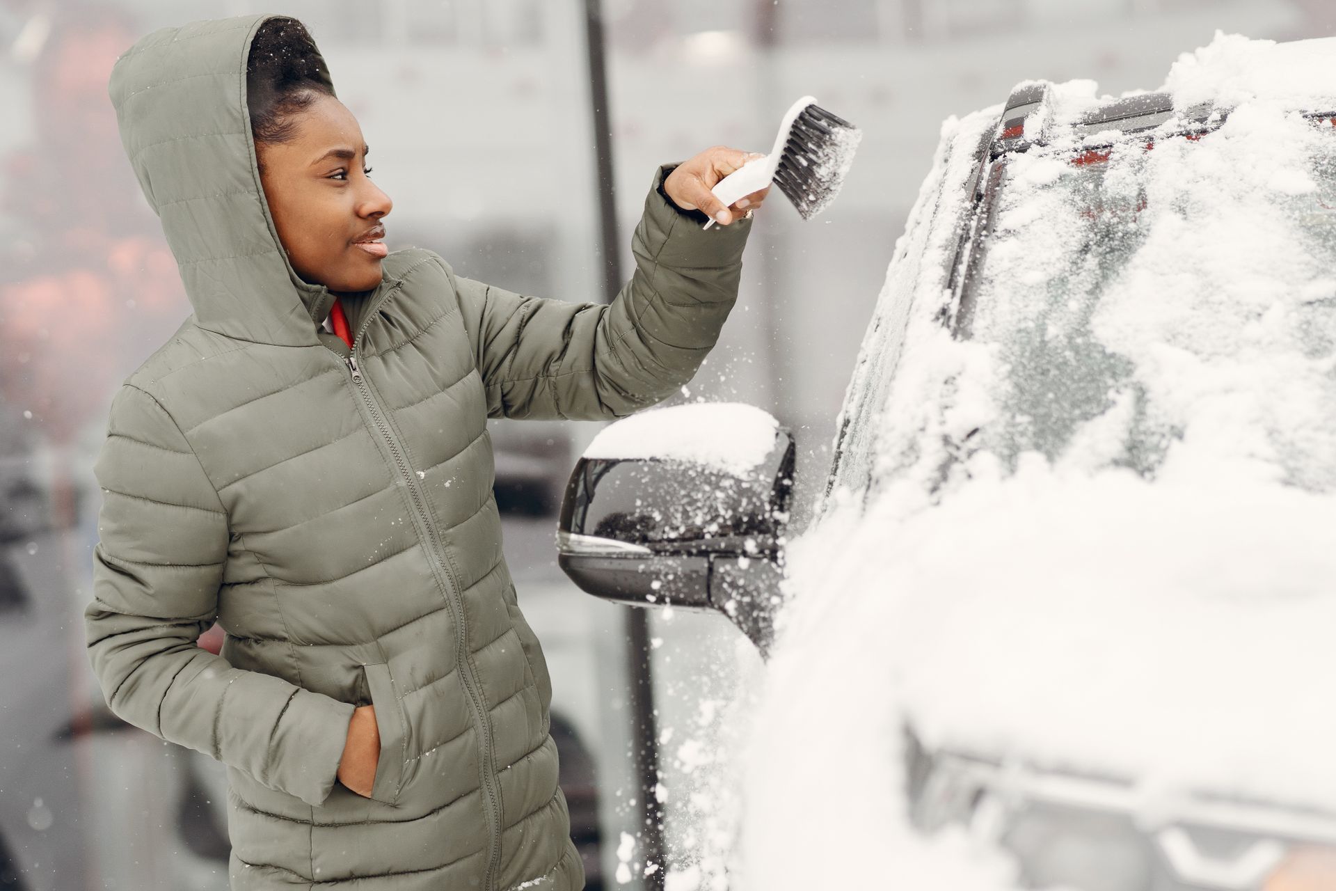 A woman in a green jacket removes snow from her car during winter outdoors.