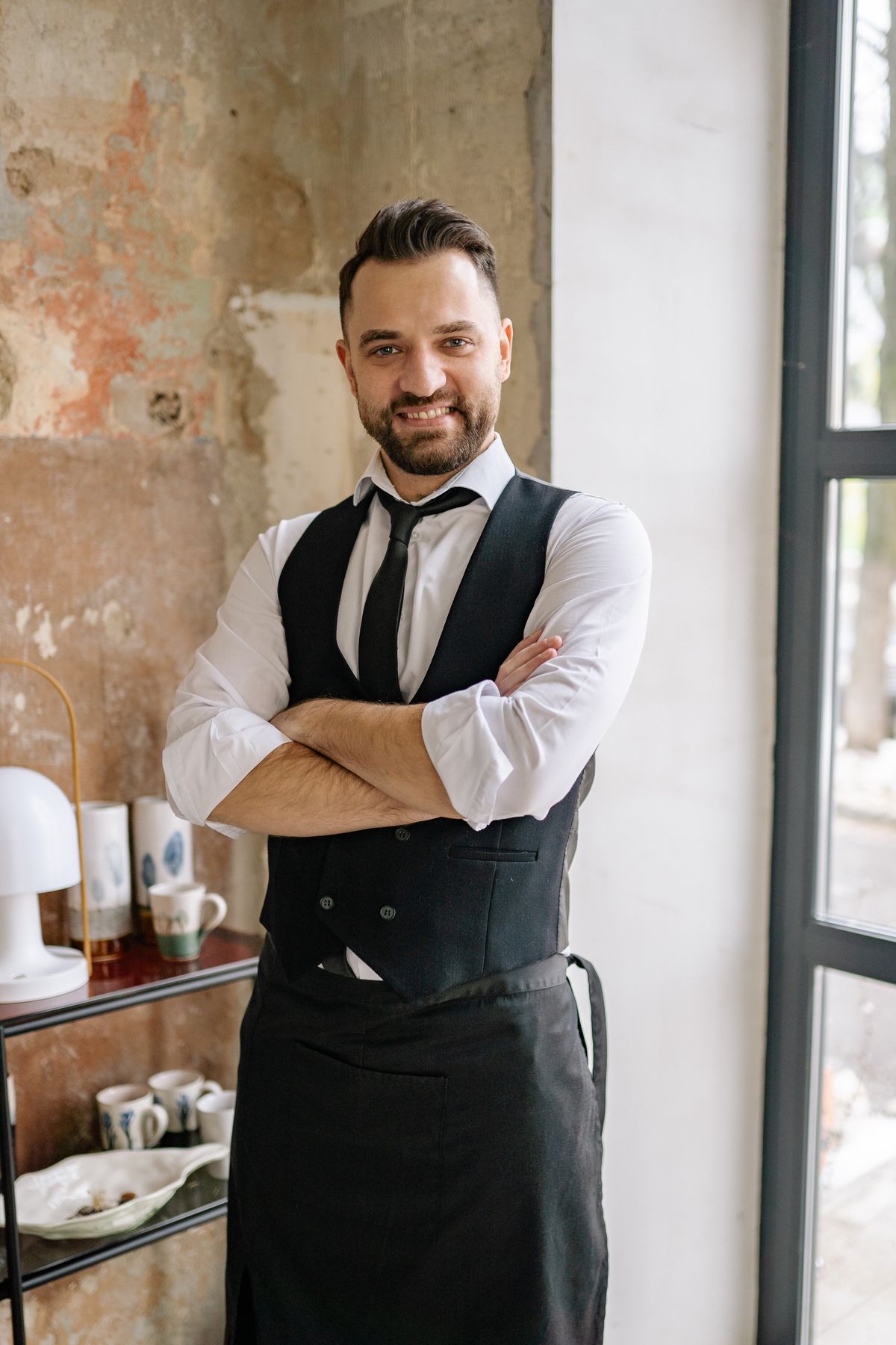 A smiling restaurant professional in a vest and apron standing beside a window