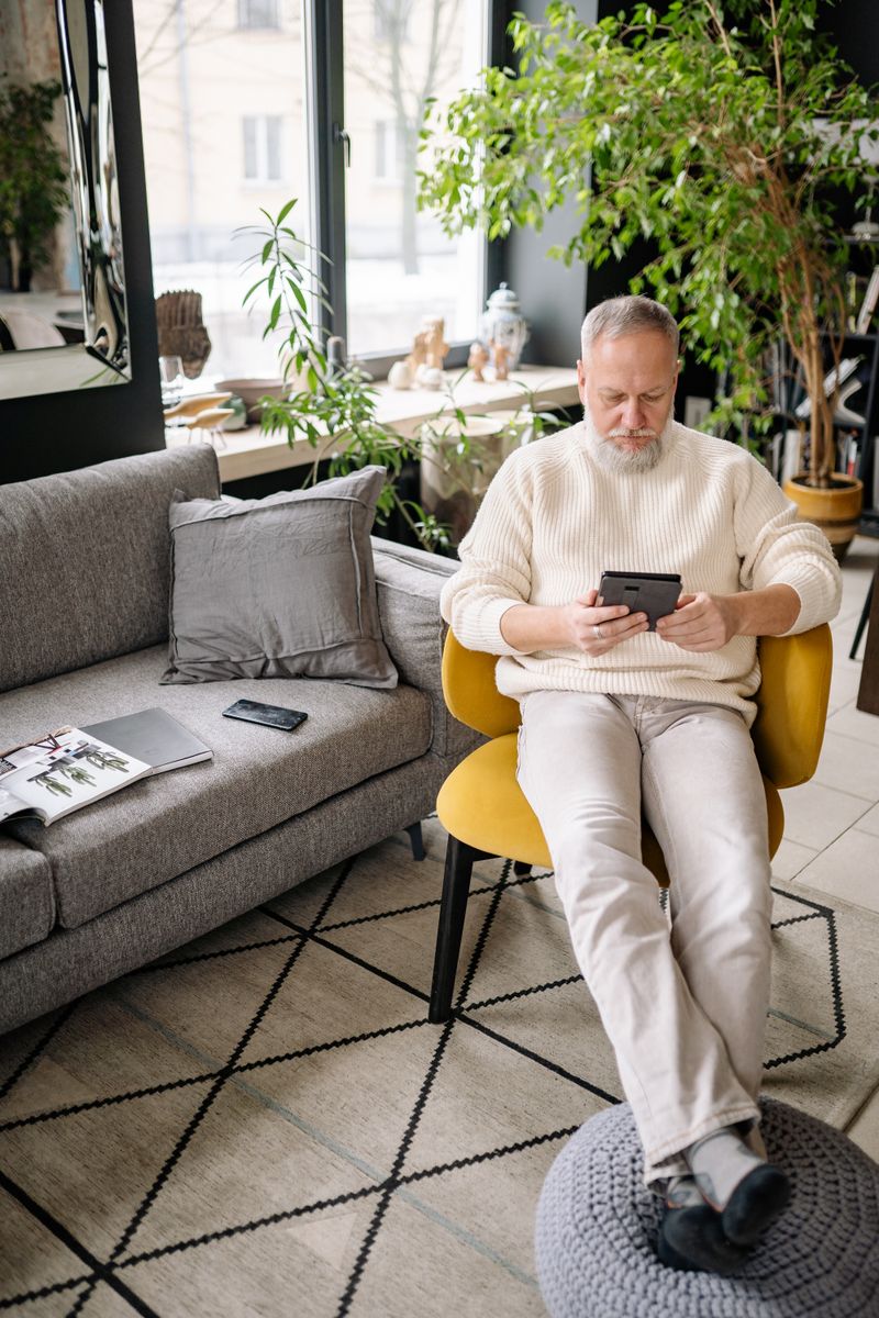 A senior man with a tablet relaxes in a cozy living room filled with plants.