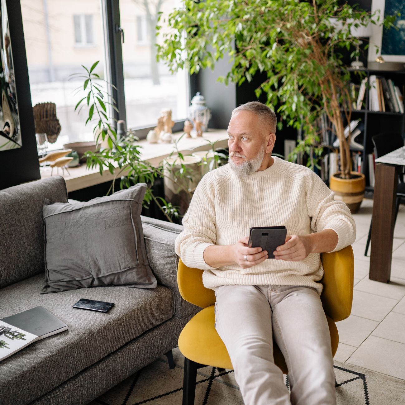 Elderly man sitting with smartphone and gadgets in a modern living room, enjoying leisure time.