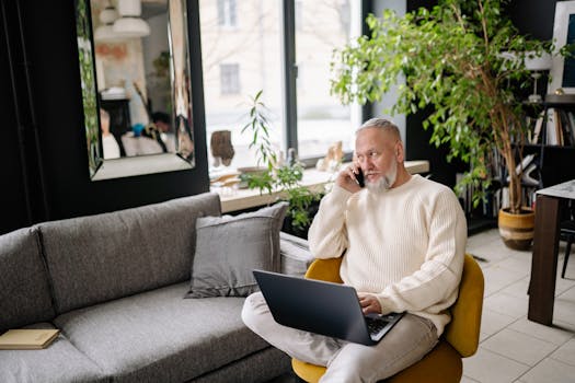 Elderly man multitasking at home, using laptop and phone, sitting comfortably indoors.