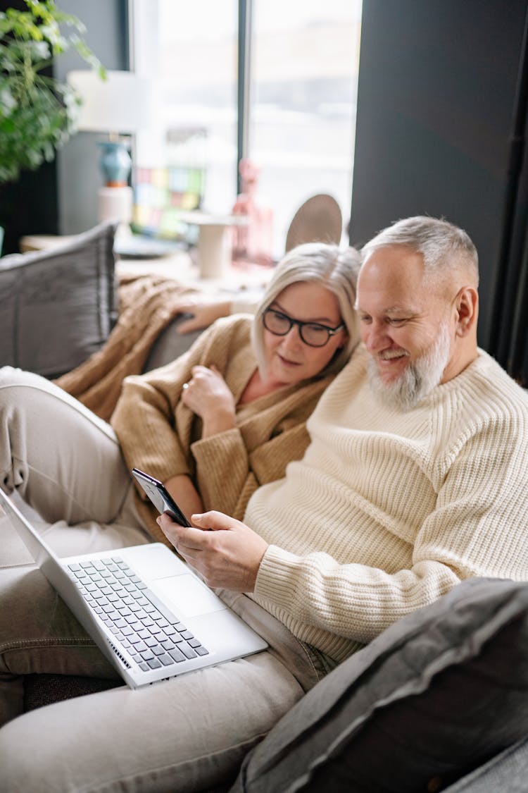 An Elderly Couple Sitting On A Couch While Looking At The Screen Of A Cellpone