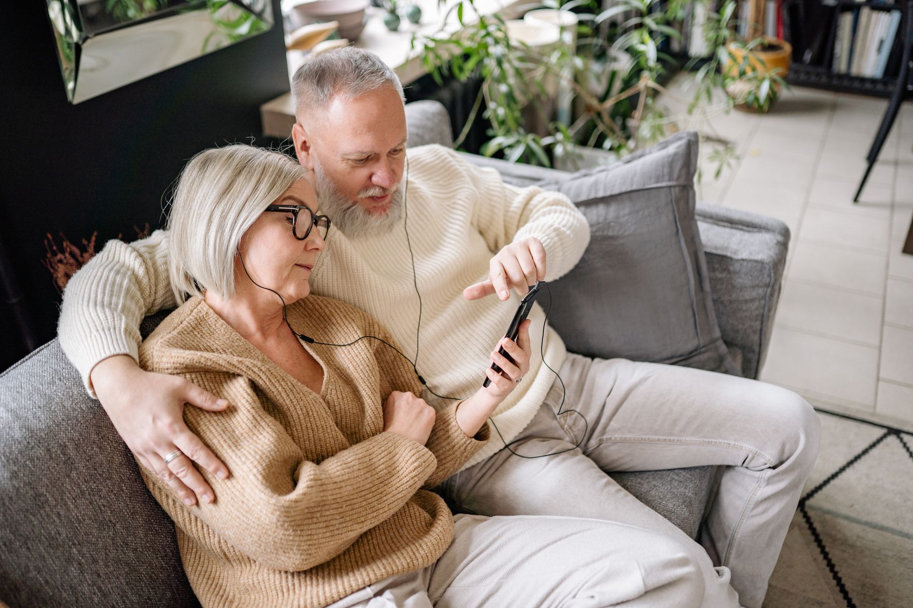 An elderly couple enjoying music on a smartphone while relaxing on a sofa indoors.