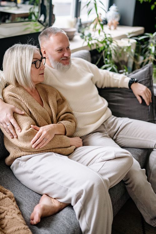 Free Senior couple sitting on a couch with an arm around each other, enjoying a relaxed moment indoors. Stock Photo