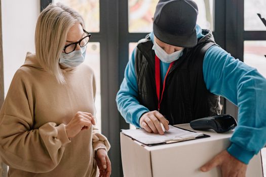 Elderly woman receives a parcel from delivery man wearing masks, highlighting contactless service indoors.