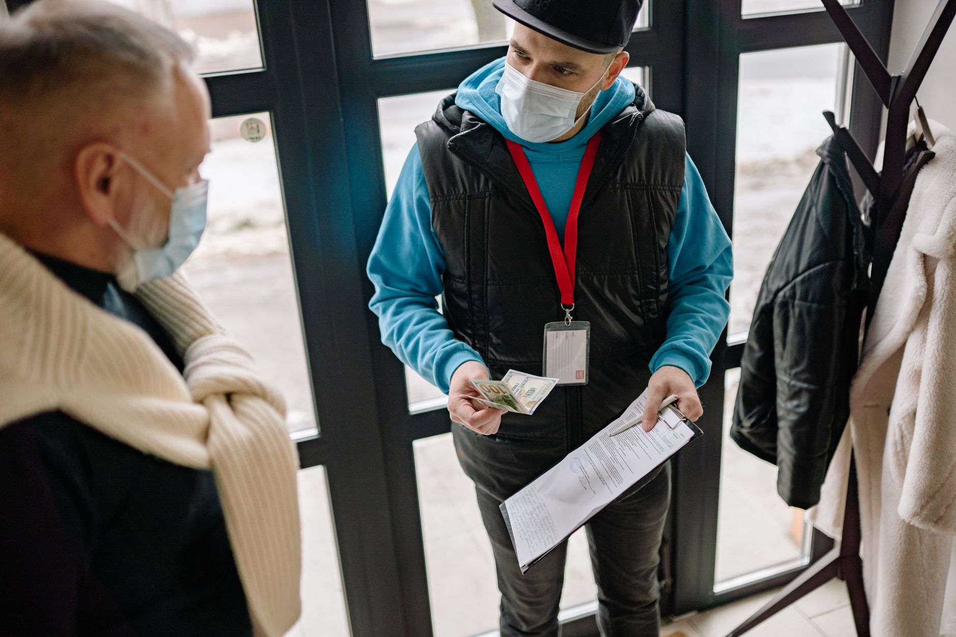 A delivery man wearing a facemask receives cash payment from an elderly man indoors.