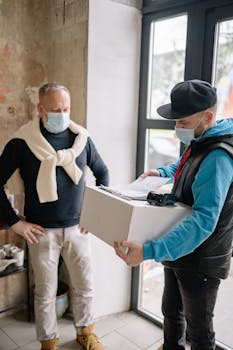 A delivery man wearing a mask hands a package to a customer inside a building.