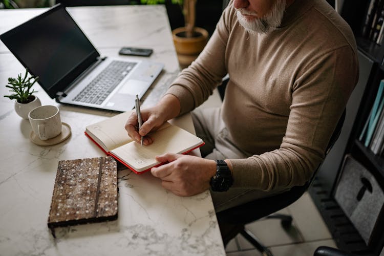 A Man In Long Sleeves Writing On A Notebook While Sitting On A Chair