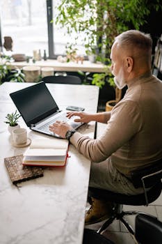 Adult man with beard working on laptop in well-lit indoor workspace with plants.