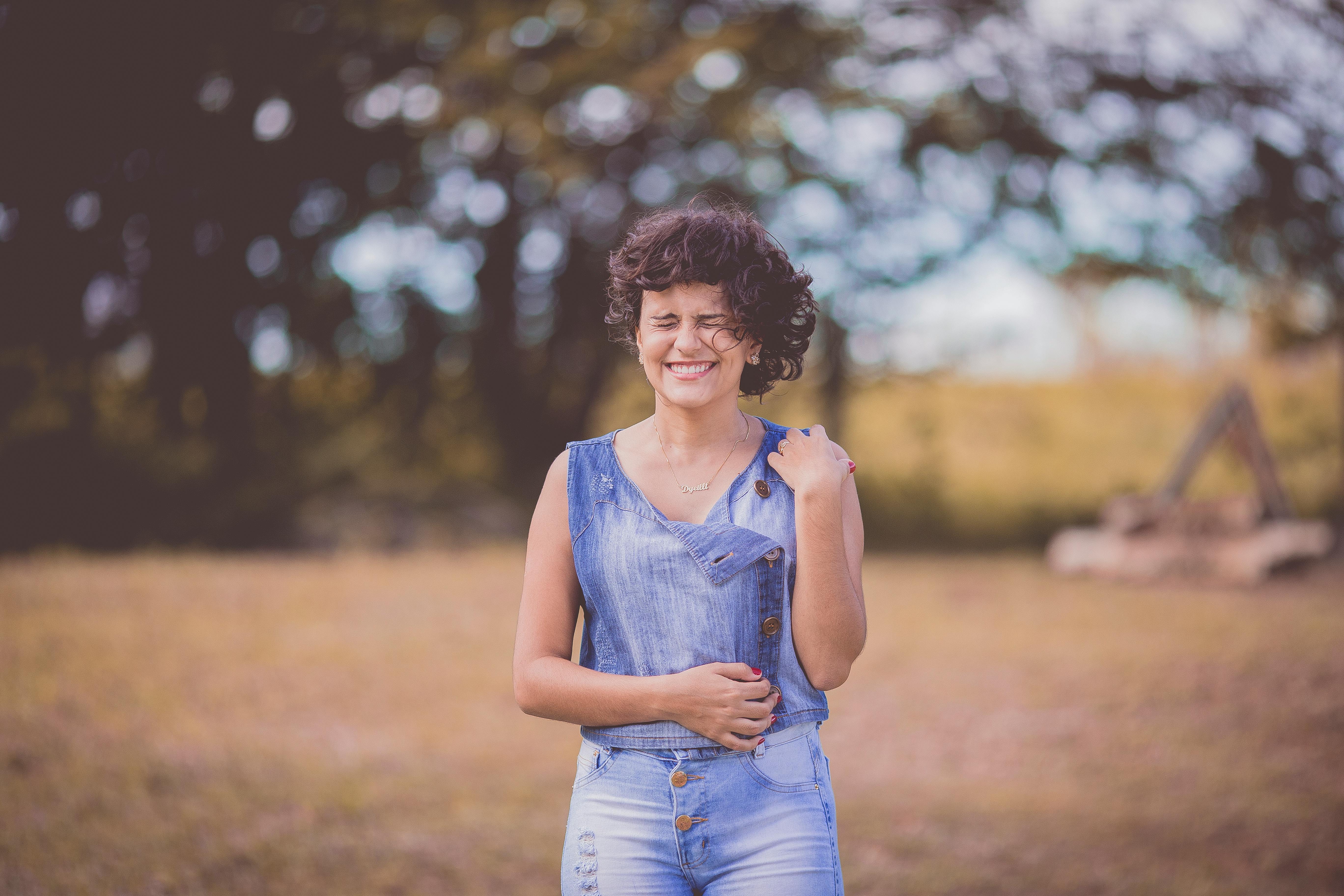 A happy woman with curly hair stands smiling in a sunny outdoor park setting.