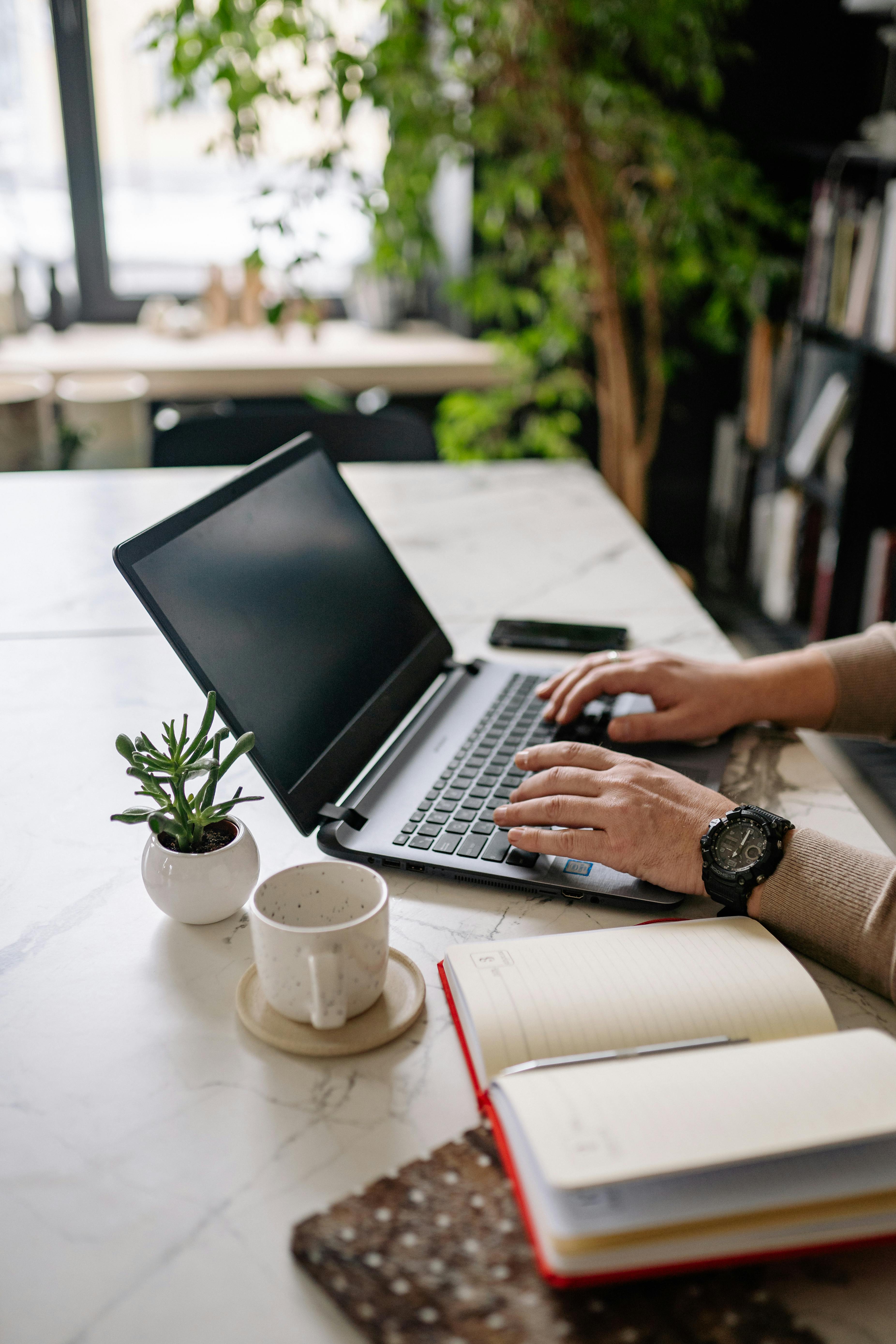 Adult typing on a laptop surrounded by indoor plants, notebooks, and a cup in a modern workspace.