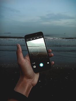 Hand holding a smartphone capturing ocean waves at sunset. Serene beach setting.