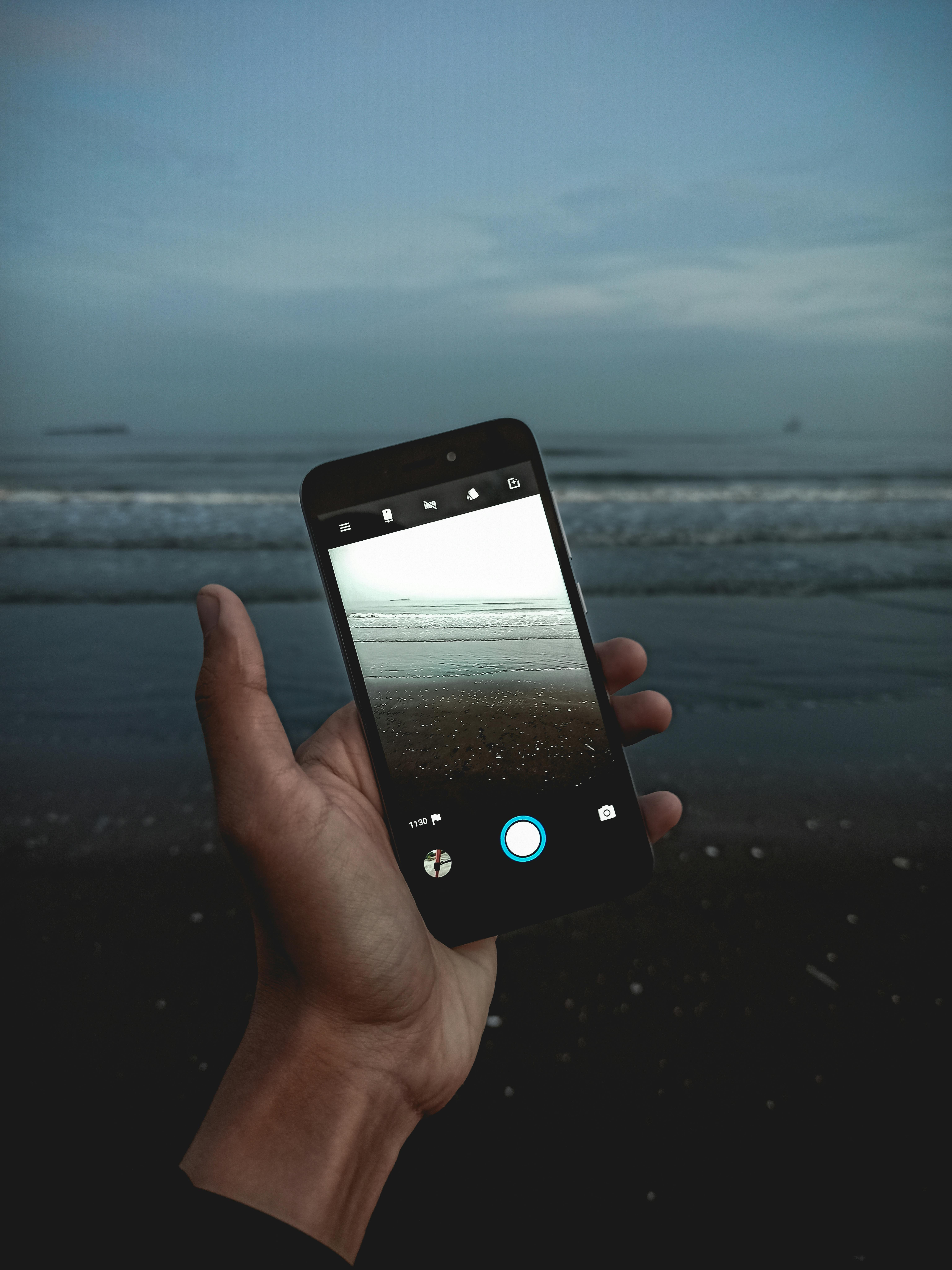 Free A hand holding a smartphone capturing the serene ocean view during evening. Stock Photo