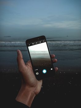 A hand holding a smartphone capturing the serene ocean view during evening.