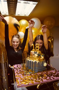 Two young women celebrating an 18th birthday party with a cake and balloons indoors.
