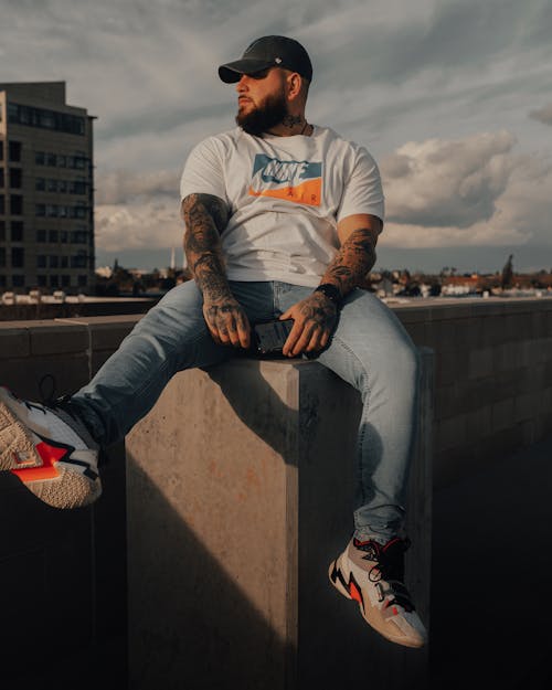 Unemotional bearded male in casual wear and cap with tattooed arms sitting on stone parapet in city district