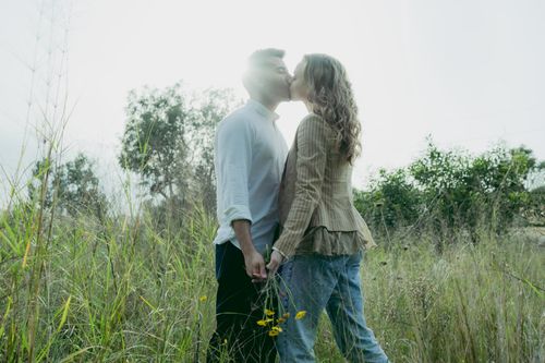 Free A couple shares a romantic kiss in a sunny meadow, surrounded by tall grass and wildflowers. Stock Photo