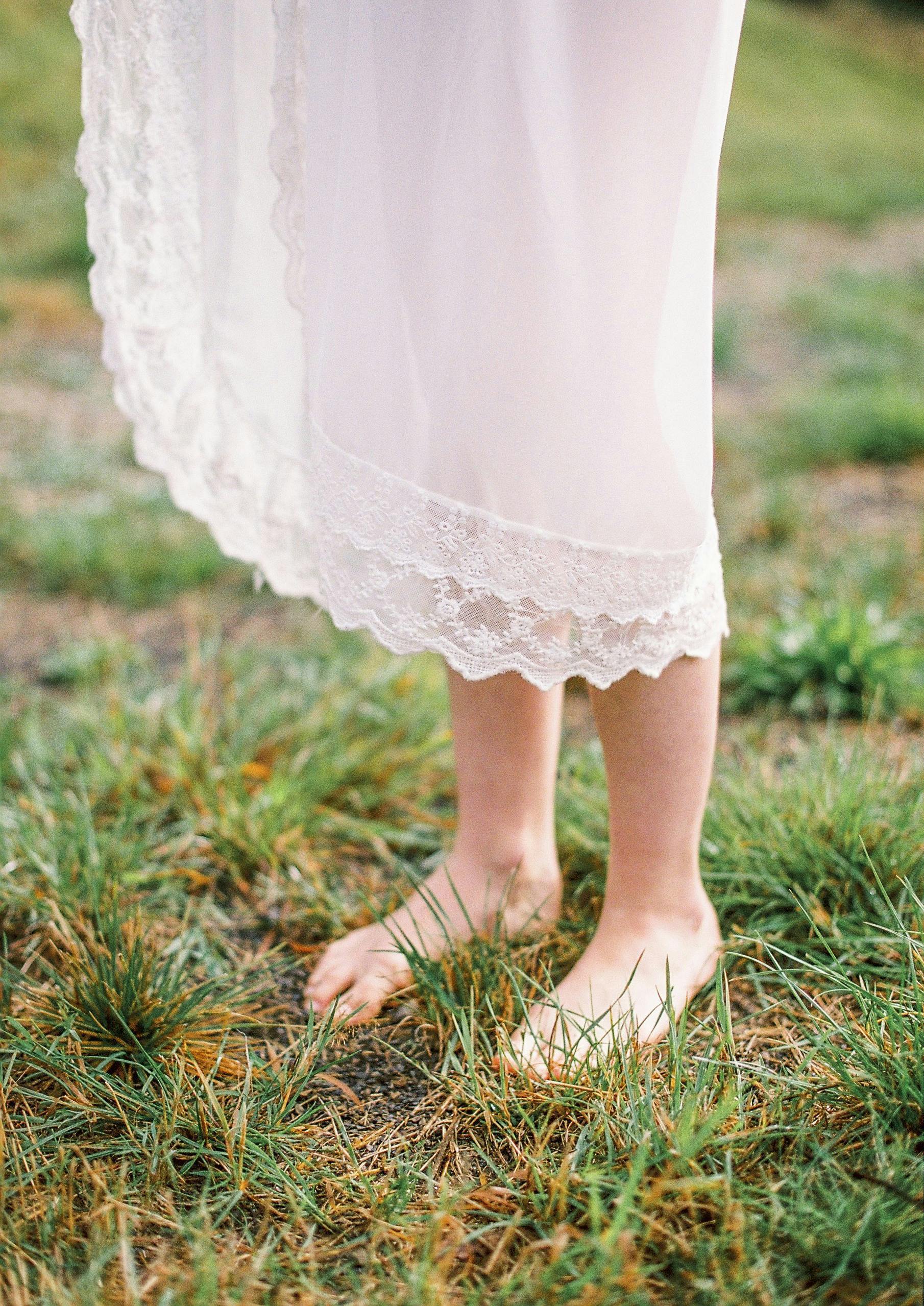 From above of crop unrecognizable barefooted female in elegant white dress standing on grassy meadow in sunny day