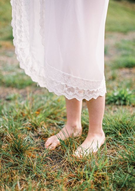 From above of crop unrecognizable barefooted female in elegant white dress standing on grassy meadow in sunny day
