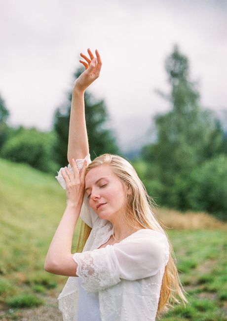 Side view of peaceful young female traveler with long blond hair standing on grassy meadow with raised arm and closed eyes while recreating in nature