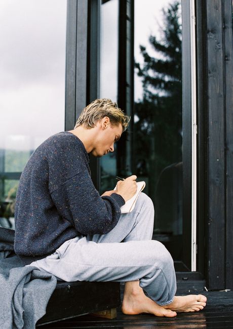 A man sitting barefoot by a window, writing in a notebook on a calm day.