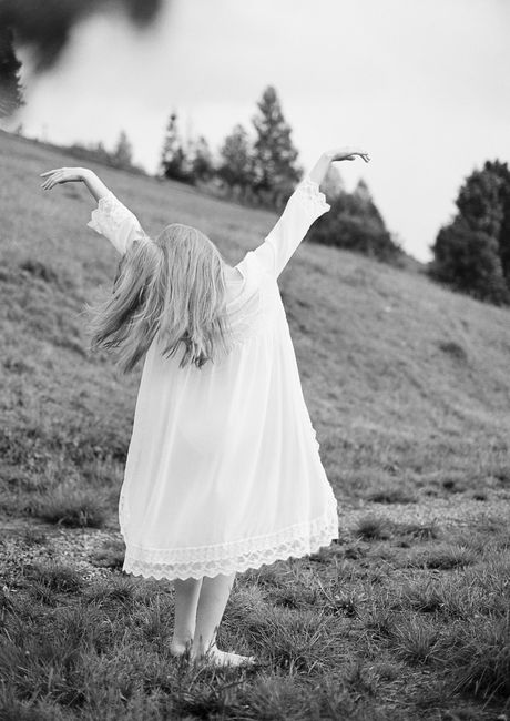 Back view of a woman in a flowing white dress with arms raised in a grassy field.