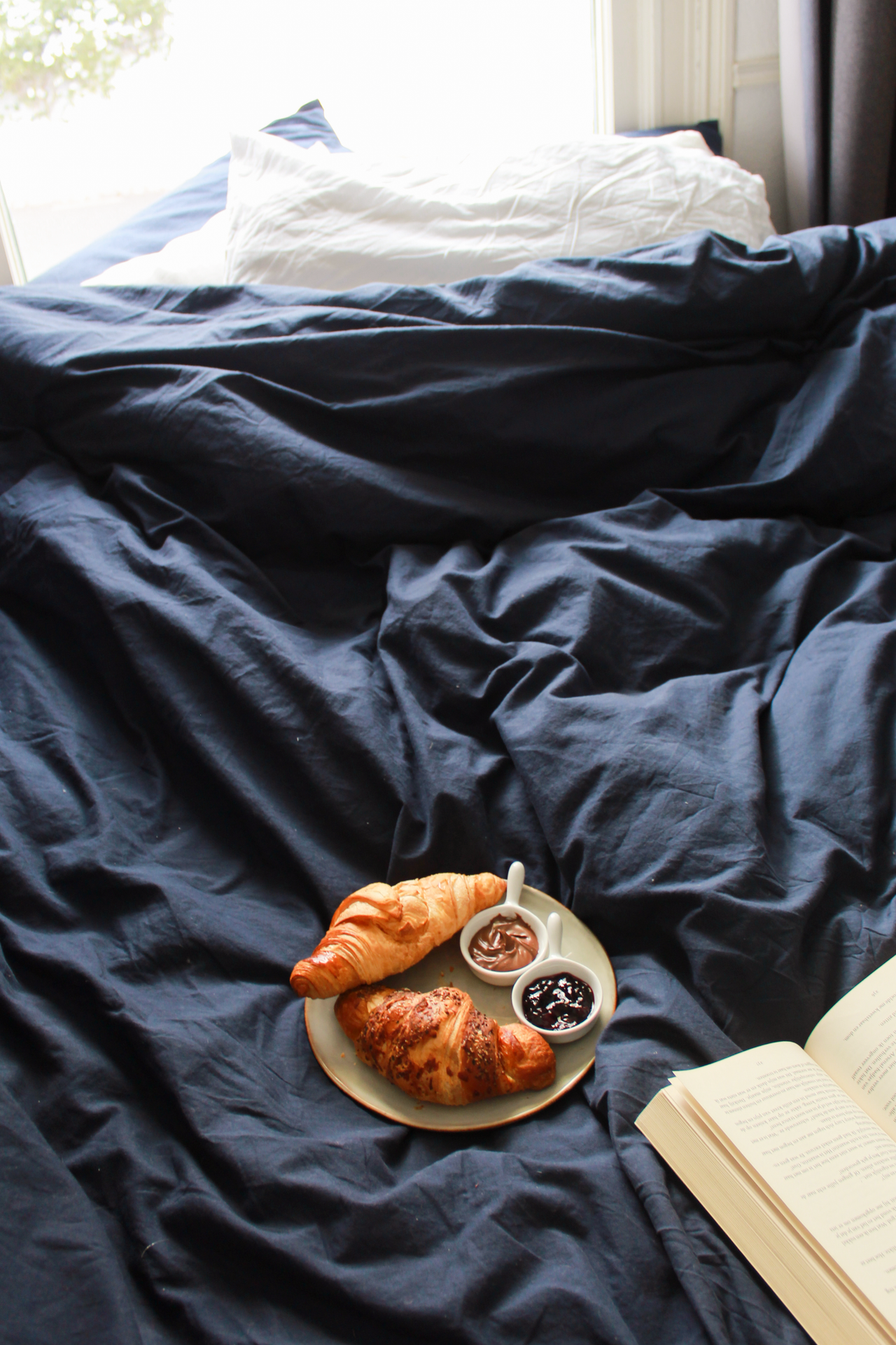 Navy blue duvet with a white pillow, a plate of croissants and an open book for a slow morning in bed