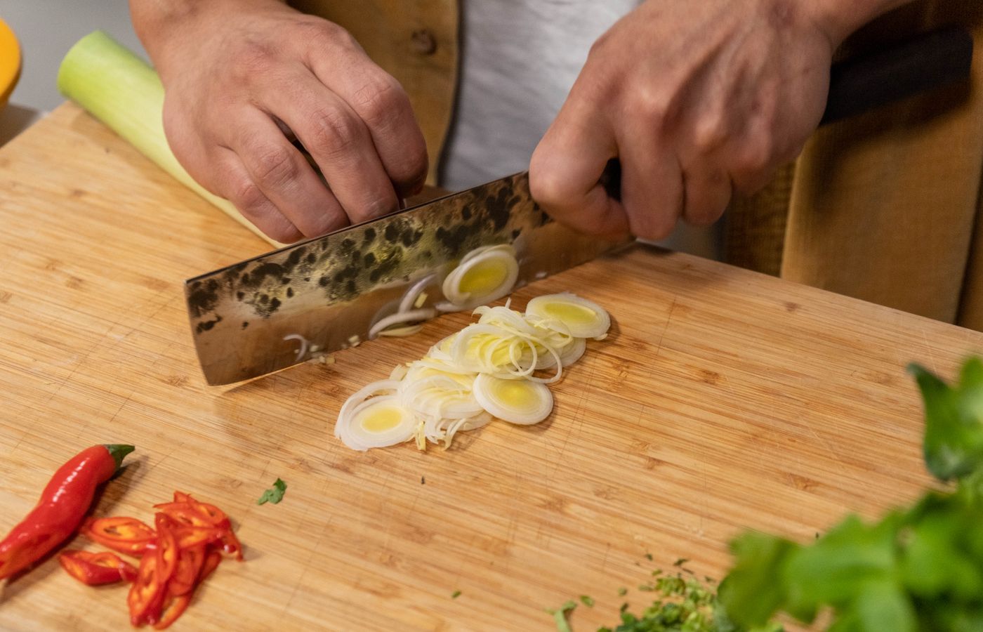 Hands chopping leeks and red chili peppers on a wooden board, showcasing culinary preparation.