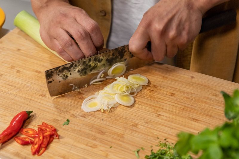 Hands chopping leeks and red chili peppers on a wooden board, showcasing culinary preparation.
