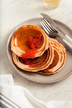 Close-up of stacked pancakes adorned with red caviar on a plate with cutlery.